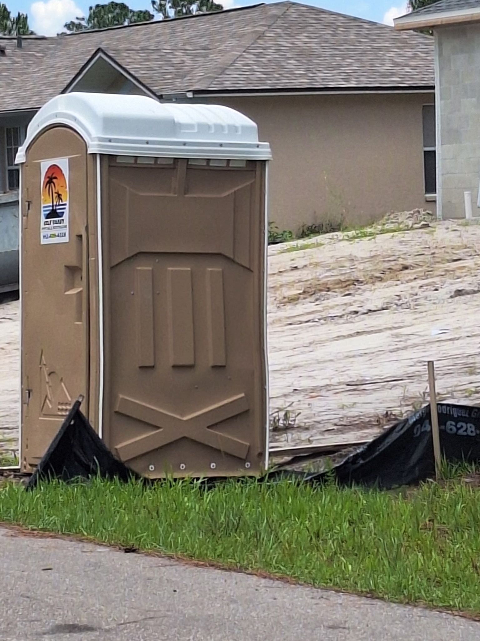Portable toilet on a dirt lot beside a road, with a house and black tarp in the background.