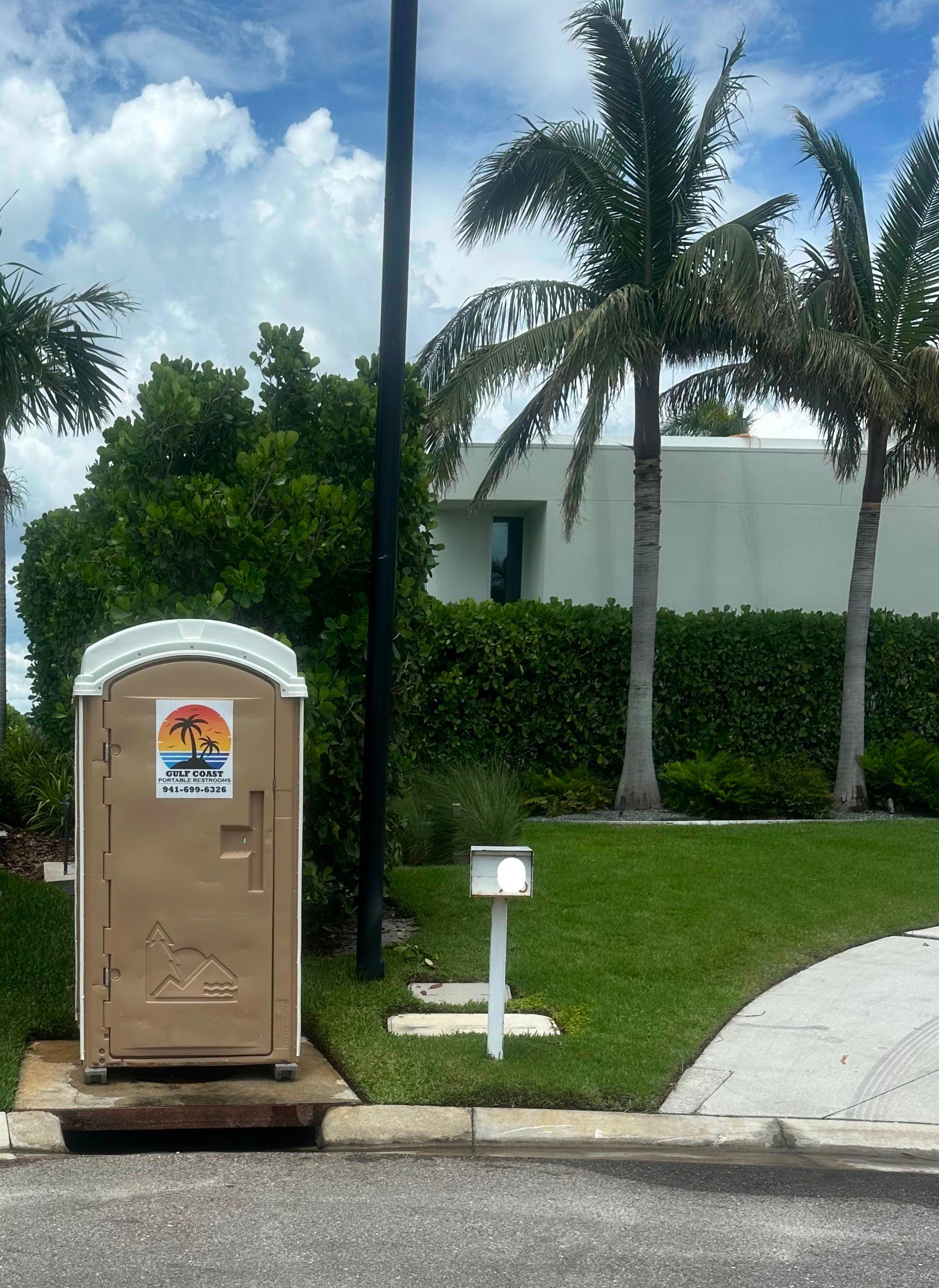 Portable toilet beside curb near palm trees and a white building on a grassy lawn