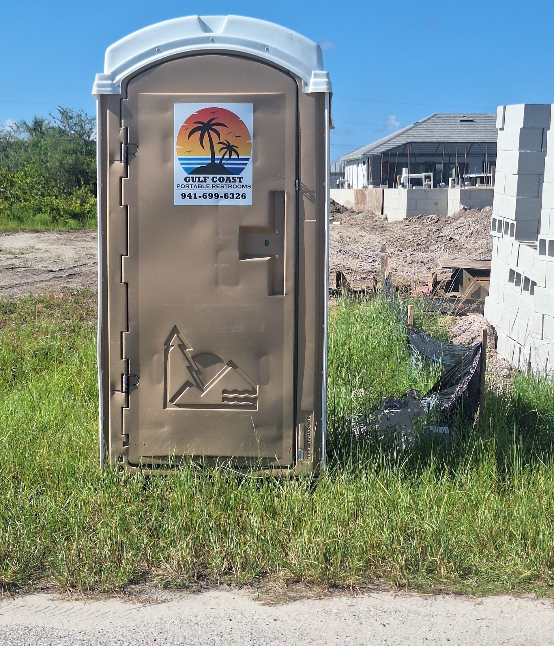 Portable toilet in a grassy lot with a building under construction in the background.