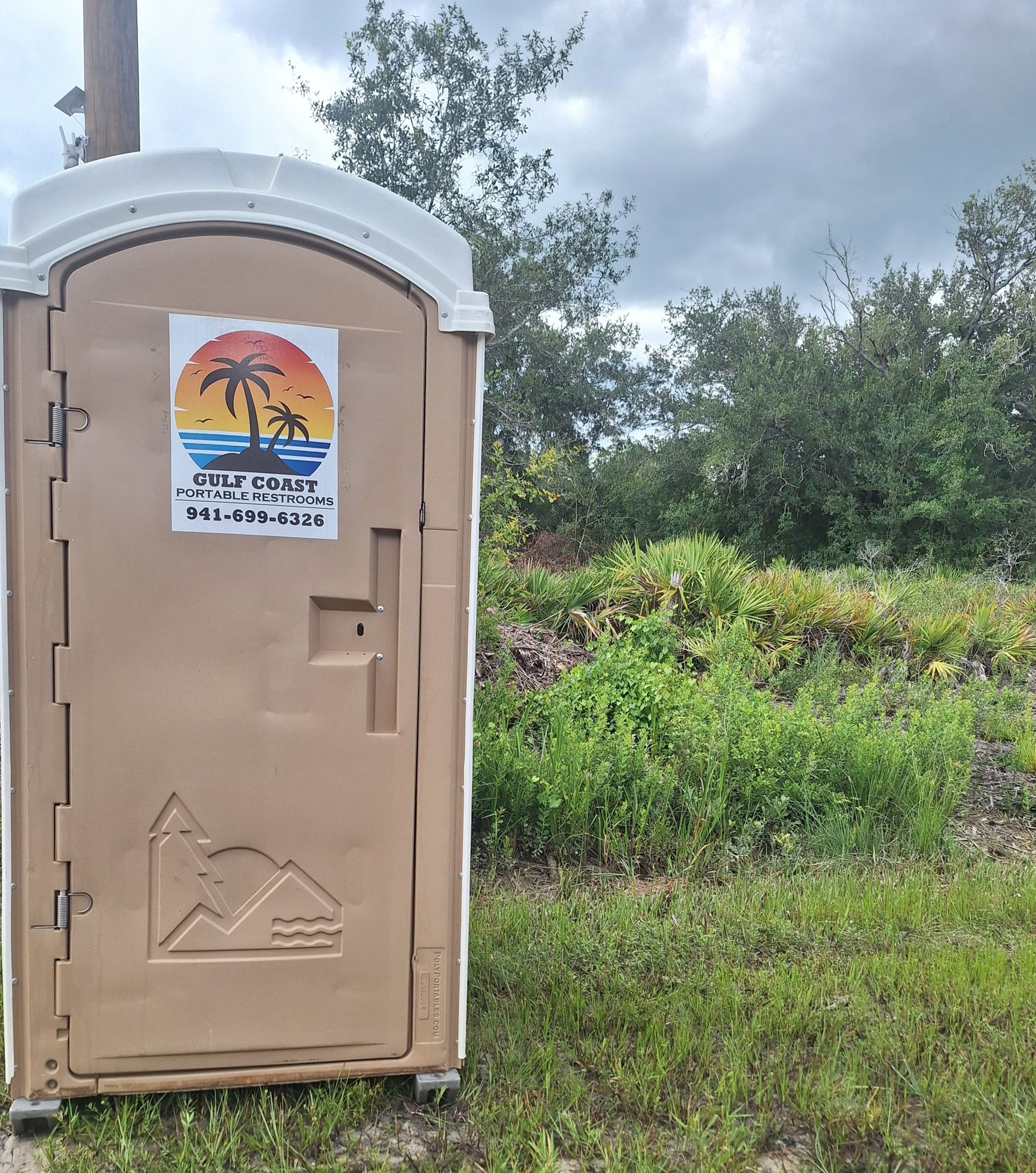 Portable toilet in a grassy field, with a sunset beach poster on the door under a cloudy sky.