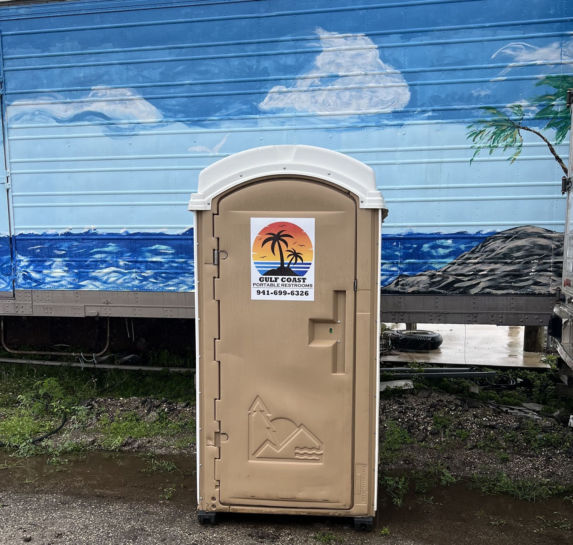 Beige portable toilet in front of a blue ocean mural with clouds and a palm tree sign.