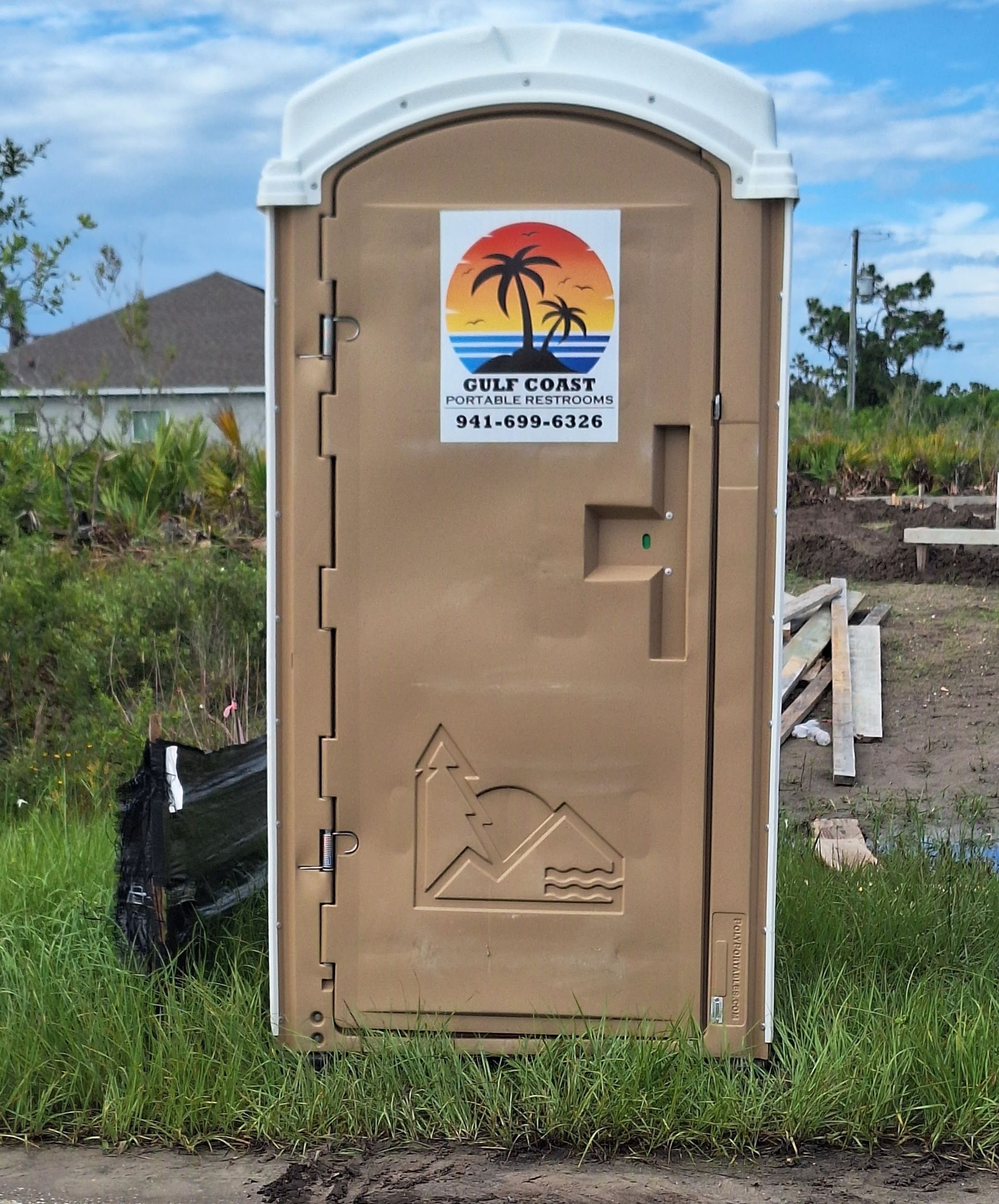 Tan portable toilet with a beach sunset logo on a grassy lot under blue sky