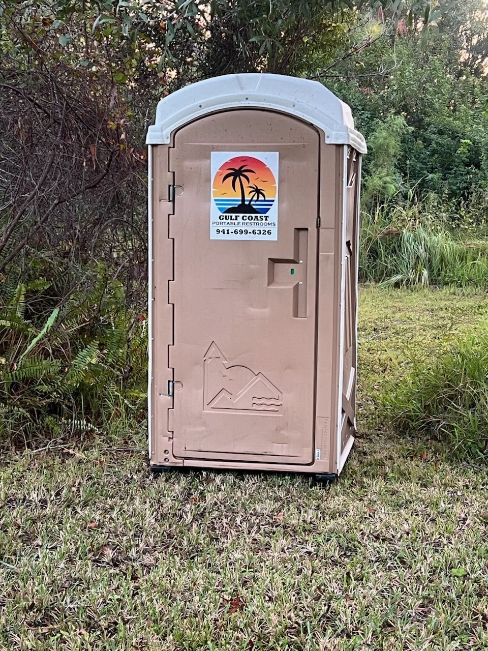 Beige portable toilet with a white roof and colorful sticker, standing on grass near trees.