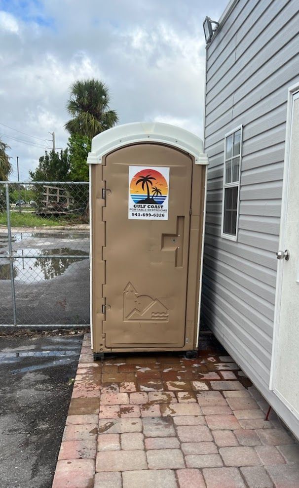 Tan portable toilet beside a gray building on wet brick pavers near a fenced lot.