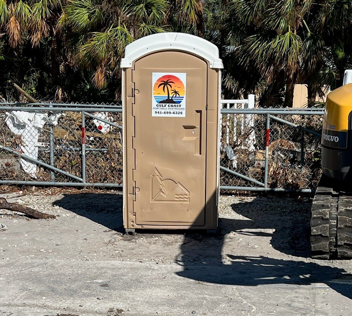 Beige portable toilet in a gravel lot with a chain-link fence and palm trees behind it