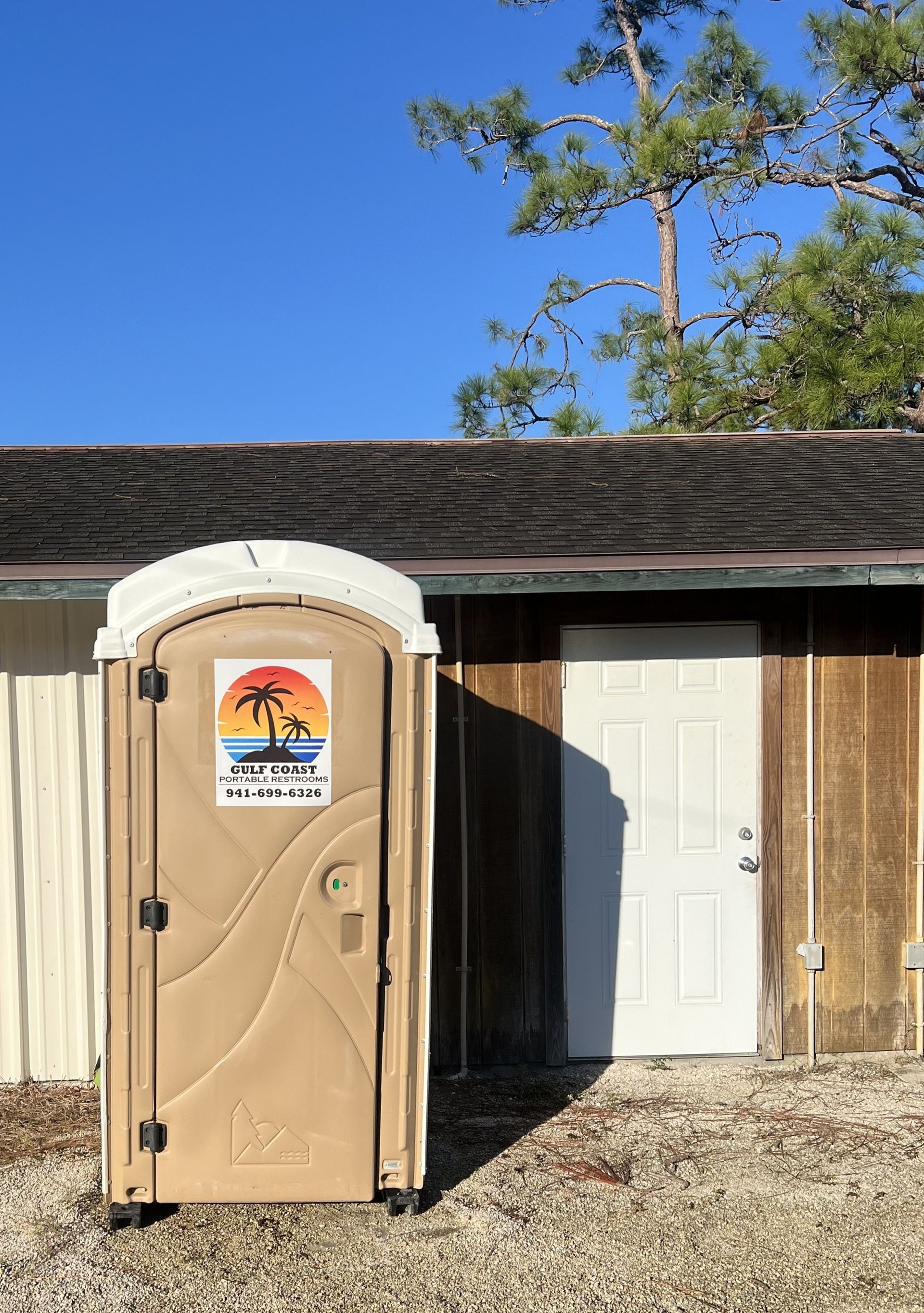 Portable toilet beside a building with a white door and a clear blue sky.