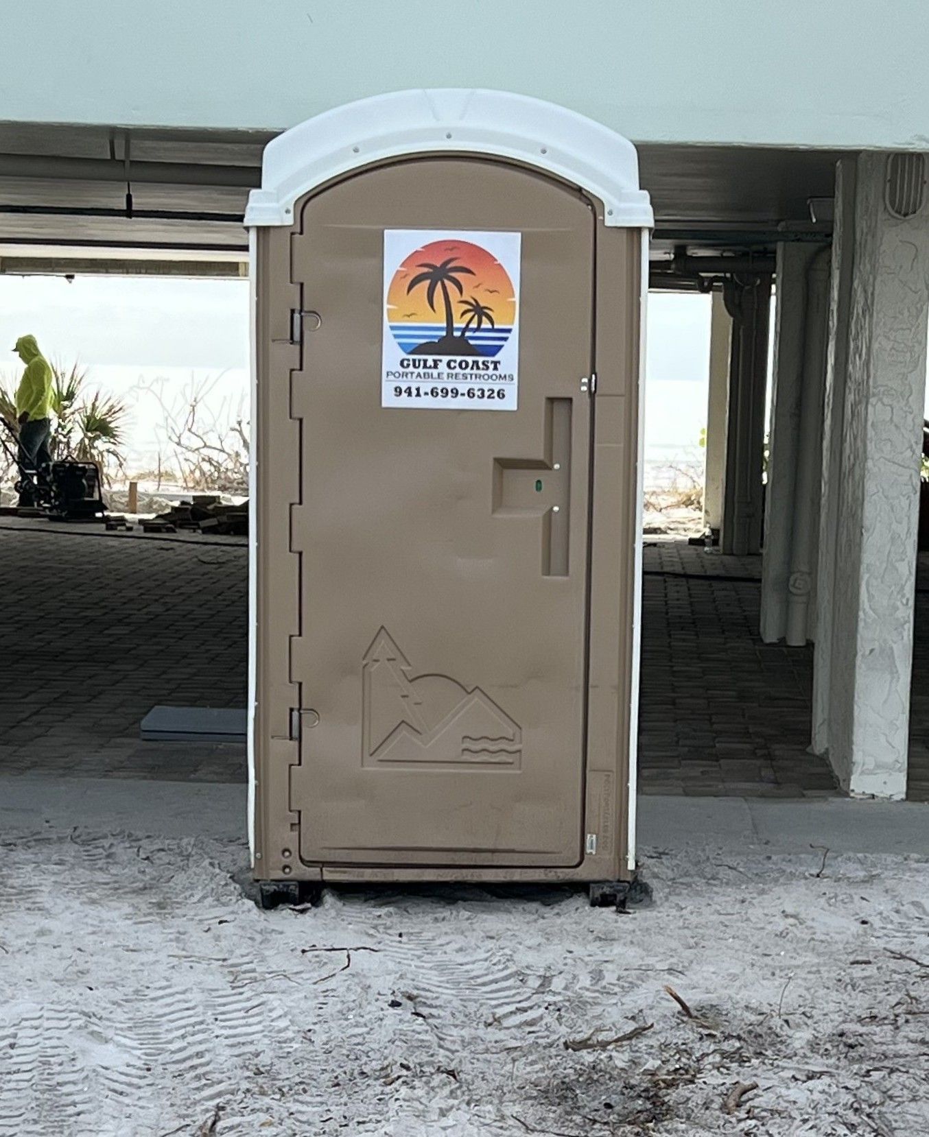 Beige portable restroom under a building, with a white roof and palm-tree sign on the door.