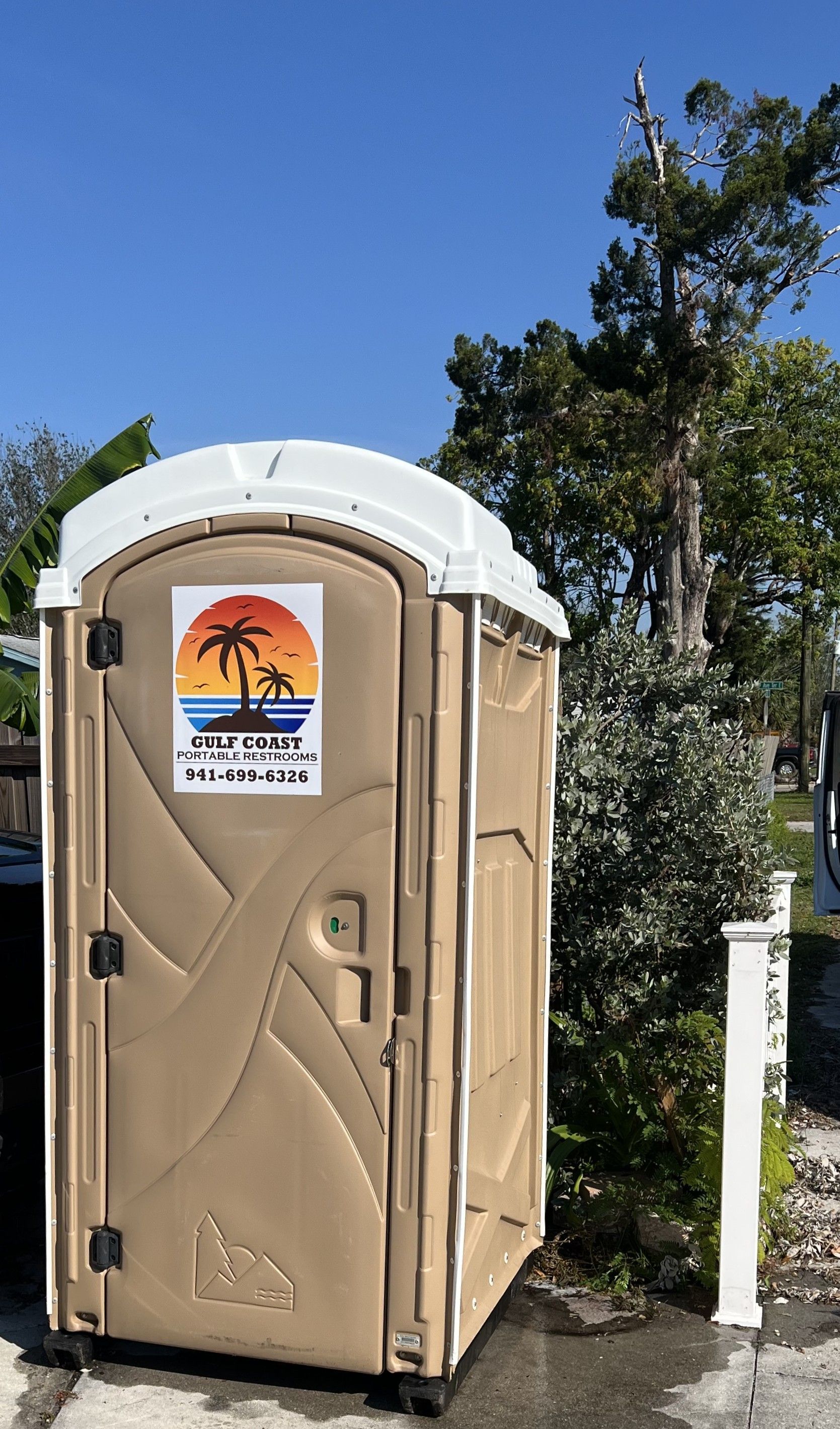 Beige portable toilet with a white roof beside a road and trees under a bright sky