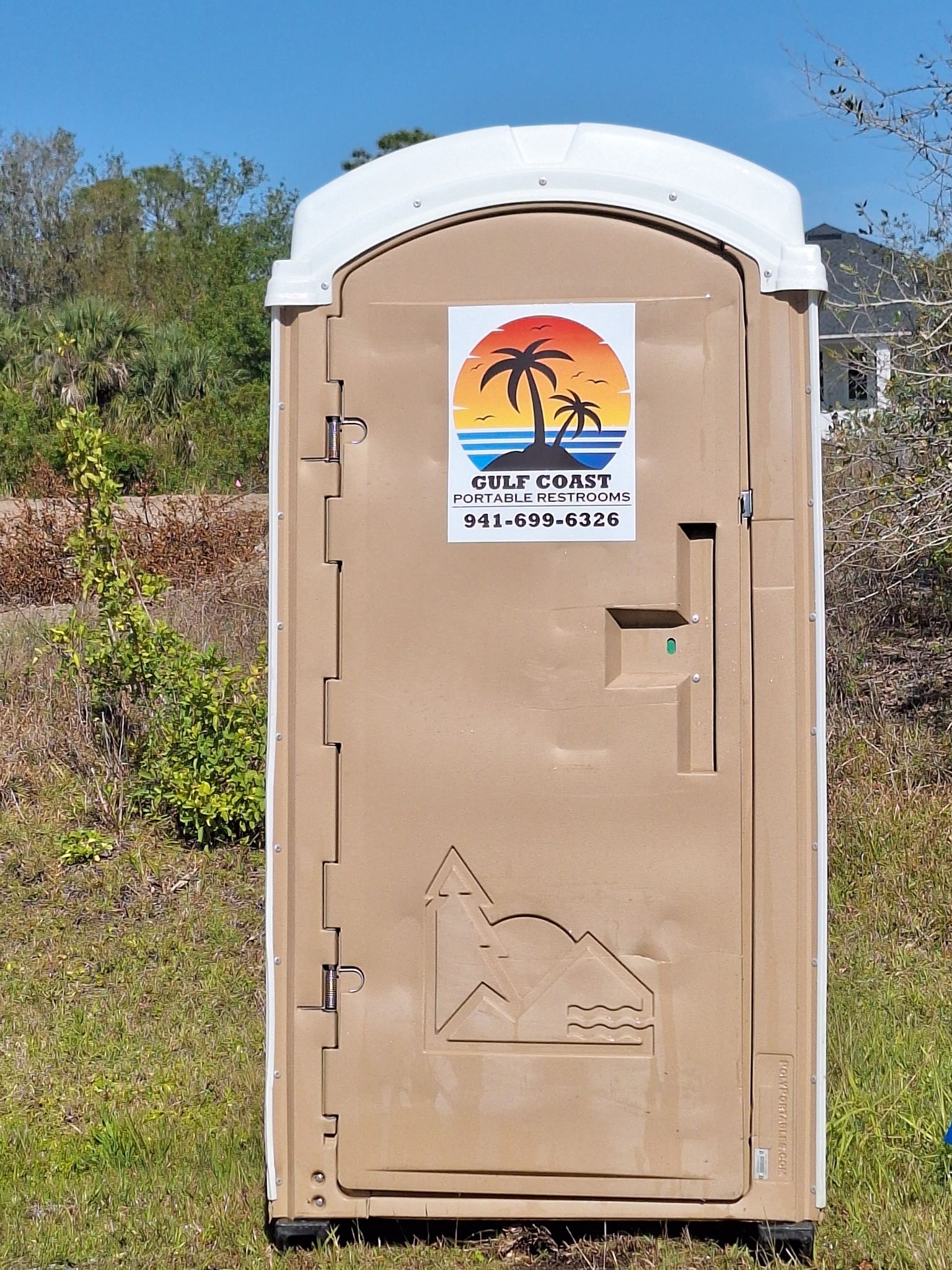 Tan portable toilet with a sunset logo sticker stands outdoors on a dirt path beside brush and trees.