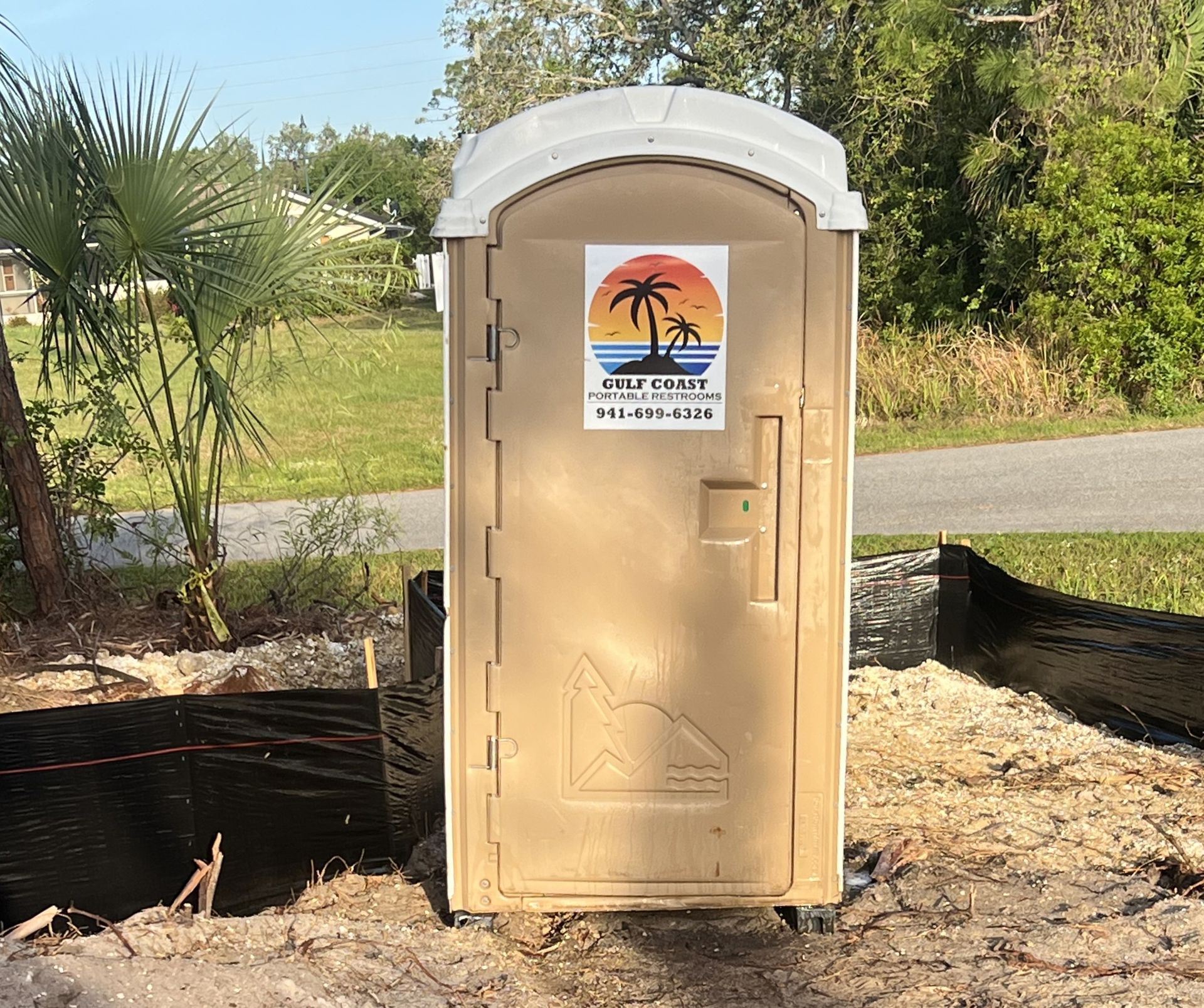 Portable beige restroom trailer with a sign, set outdoors near trees and grass.