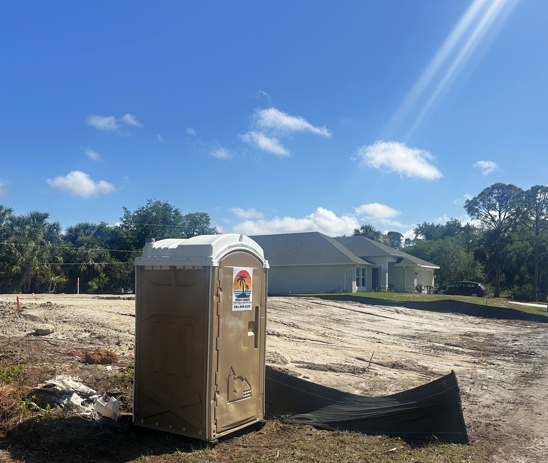 Portable toilet at a construction site under a bright blue sky, with houses in the background.