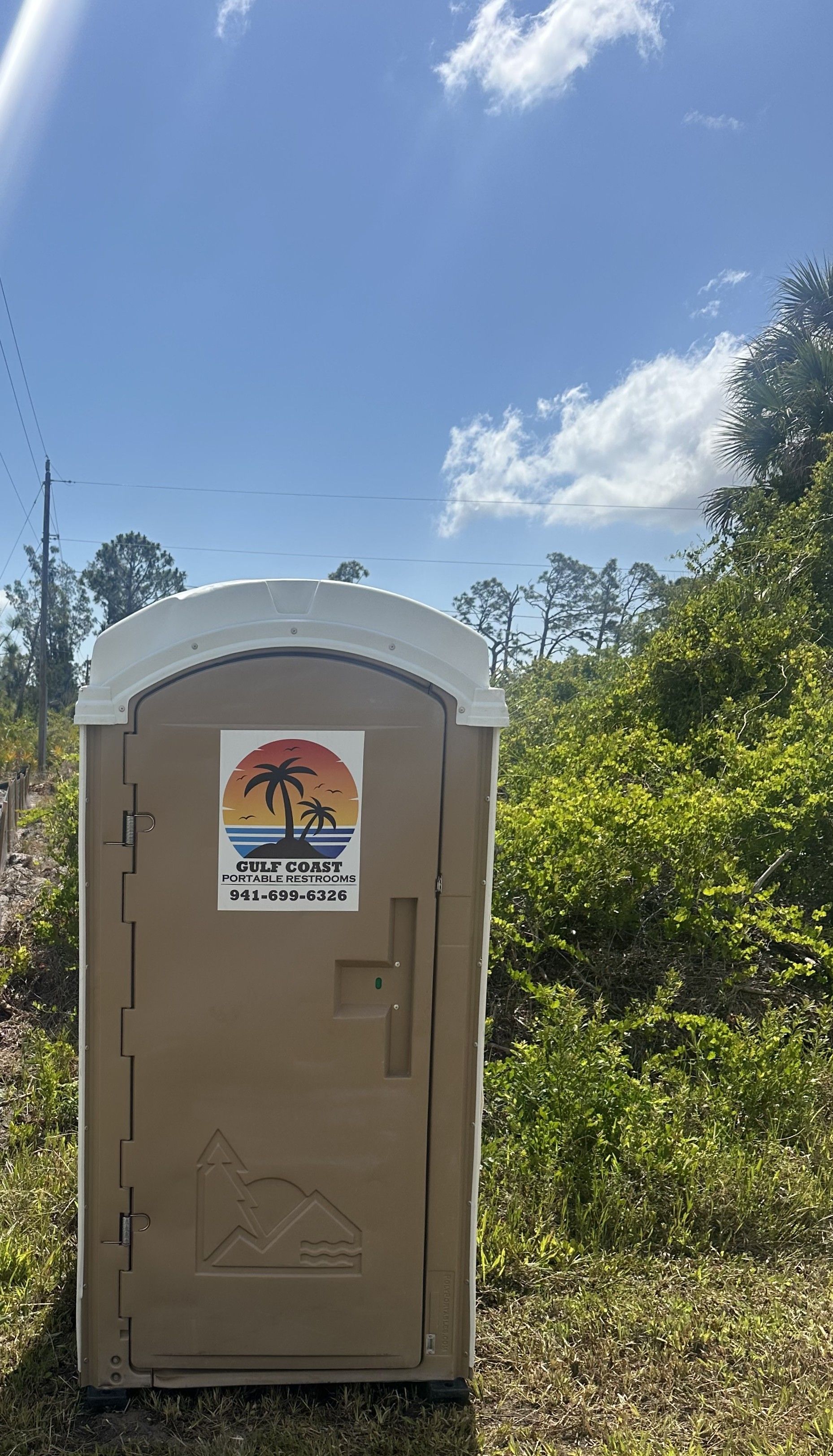 Portable toilet in a sunny outdoor area with greenery and blue sky.