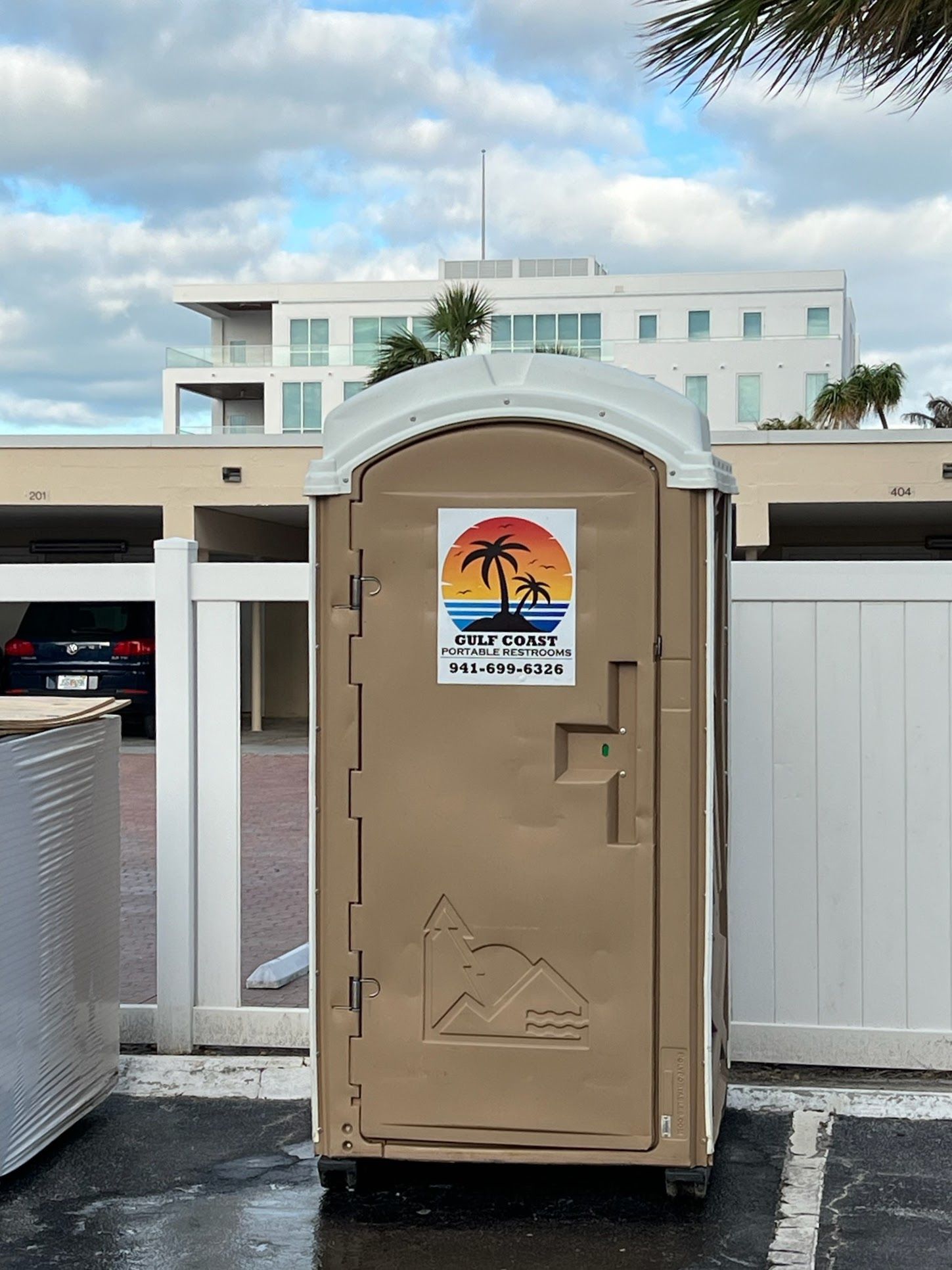 Portable toilet in front of a white building, with a beach sign on the door.