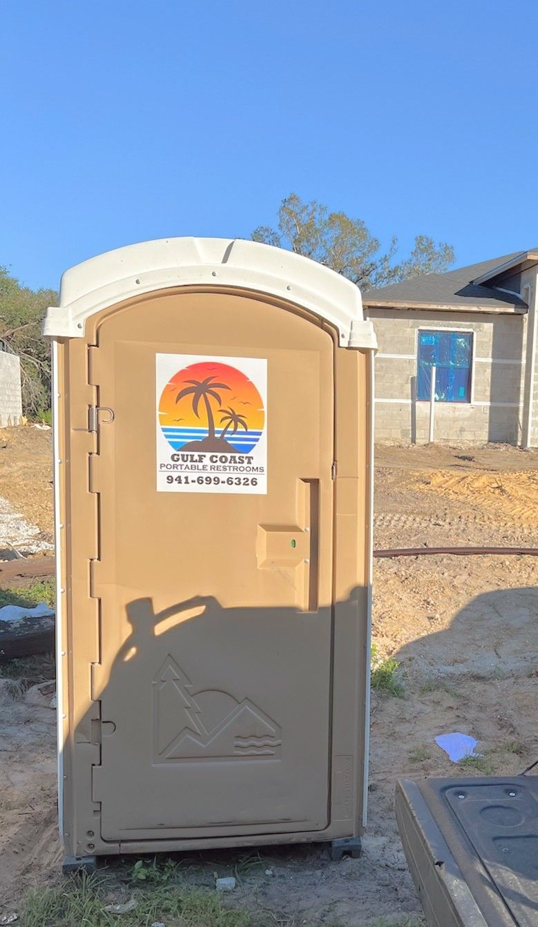 Tan portable toilet with a palm tree sticker in a dry outdoor lot under a blue sky