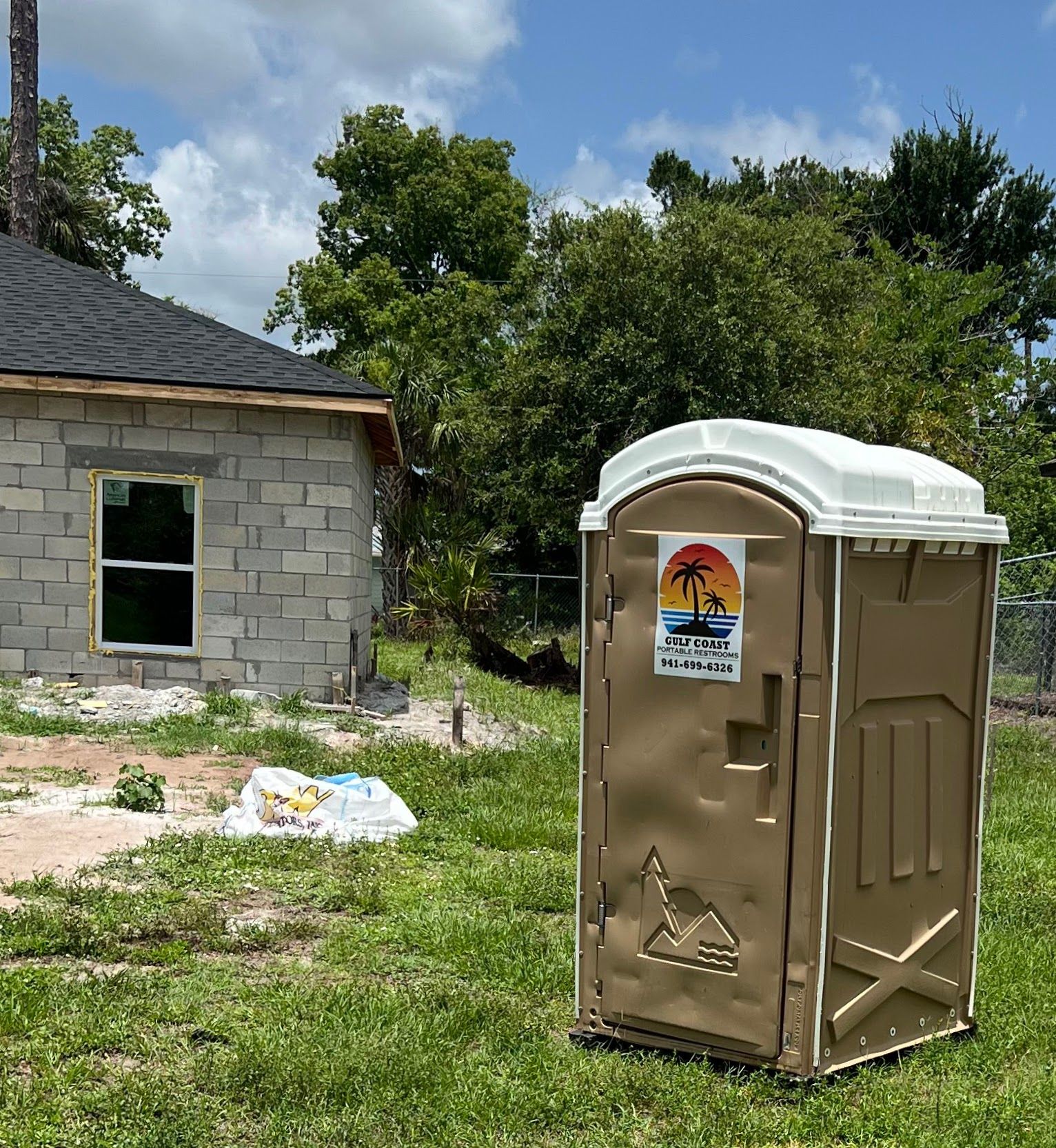 Portable toilet in a grassy yard beside a partially built cinder block house under a blue sky