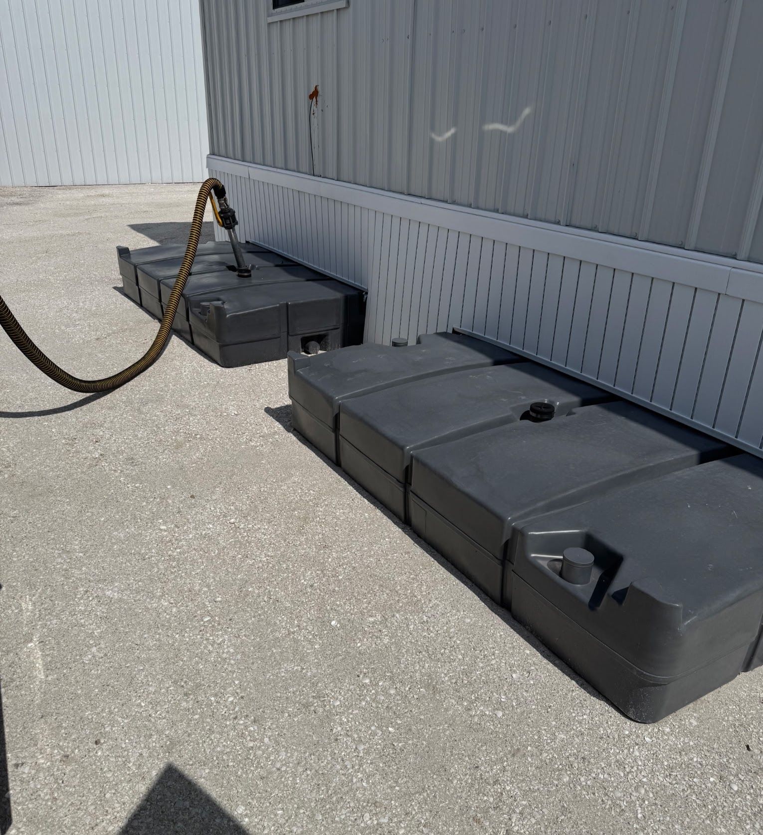 Two black utility boxes chained to a gray industrial building on a sunny pavement.