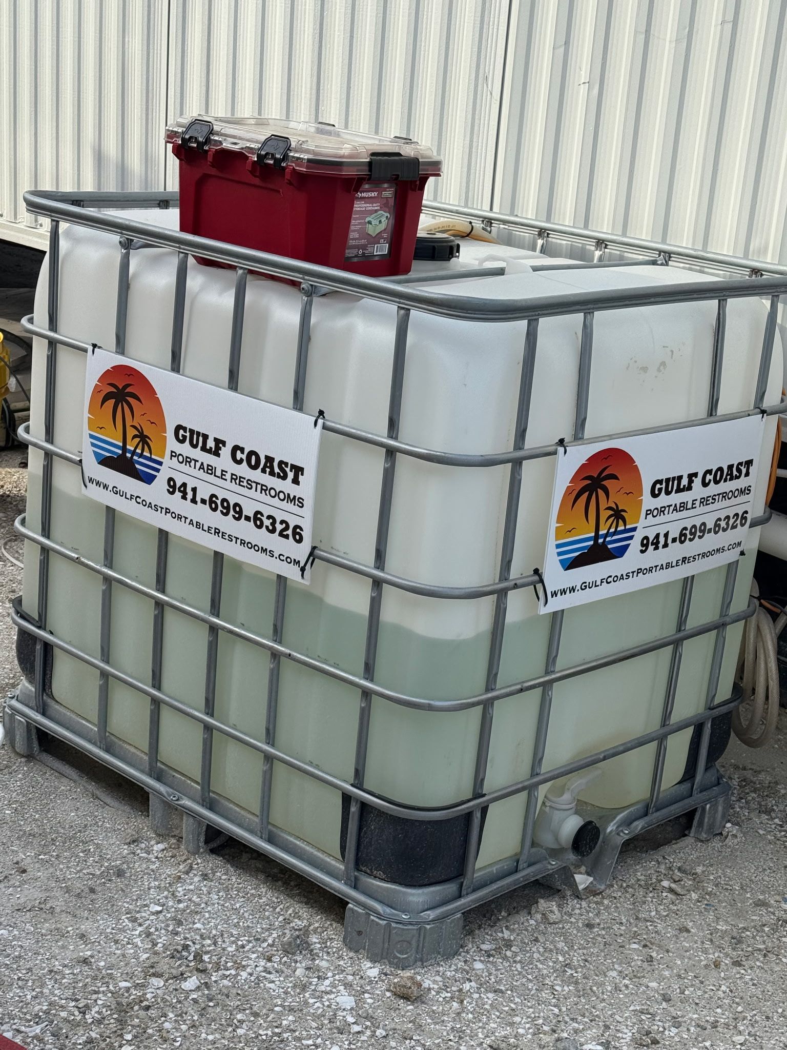 Large white chemical tote with red caps and company labels beside a corrugated metal wall