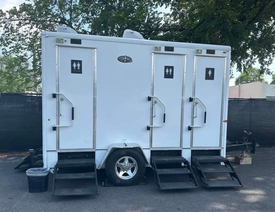 White portable restroom trailer with three doors and steps, parked outdoors near trees and fencing.