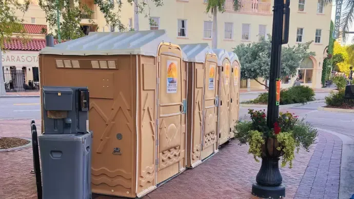 Portable restroom stalls beside a trash bin on a brick sidewalk in a town square