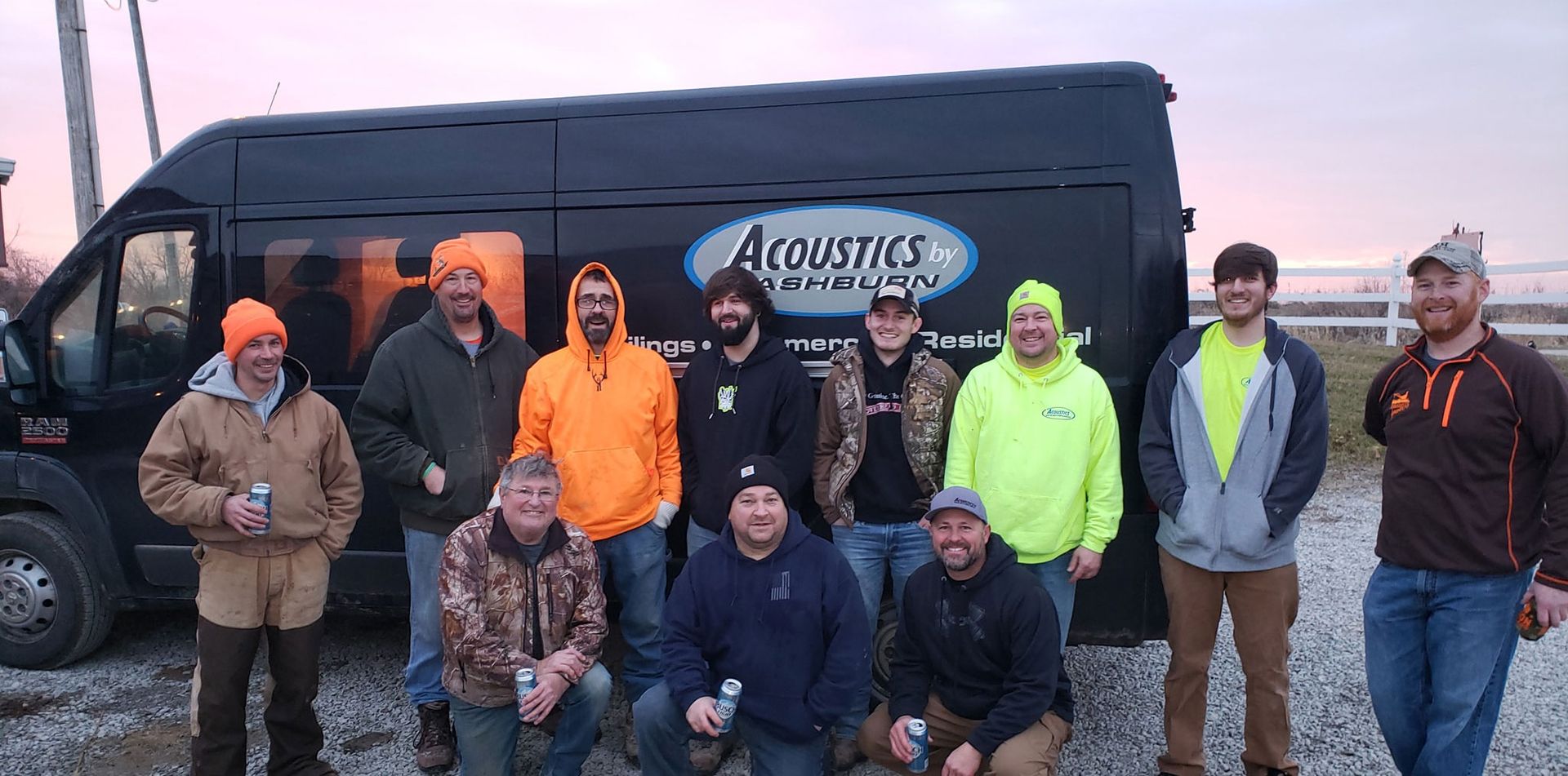 A group of men standing in front of a van with "Acoustics Equipment" logo. Some wear orange and neon. Cloudy day.