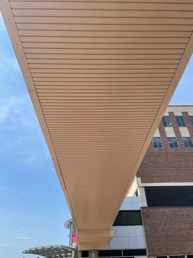 Low-angle view of a tan-colored, slatted overhead walkway. A building and sky are visible in the background.