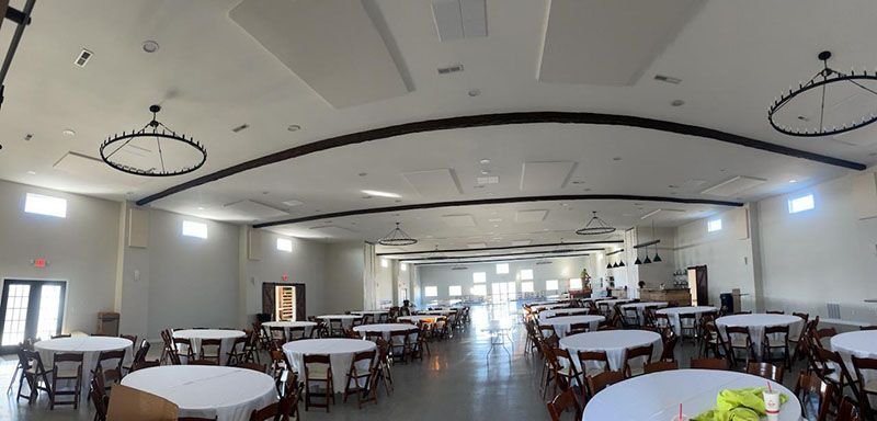 Large event space with round tables, chairs, and chandeliers. Arched ceiling with recessed lighting.