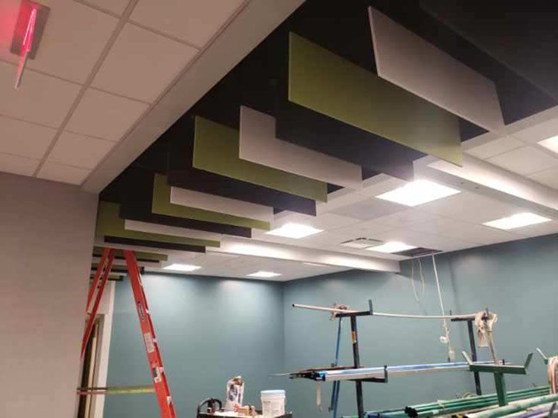 Interior view of a room with green, white, and black rectangular ceiling panels. A ladder and other equipment are visible.