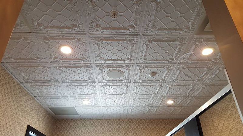 White, patterned ceiling tiles with recessed lighting and a speaker, in a room with textured walls.
