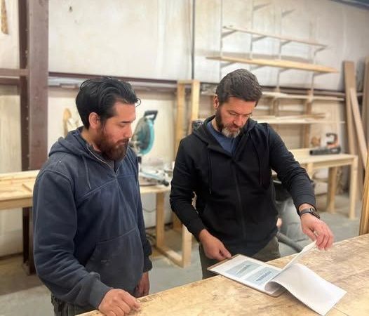 Two woodworkers in a workshop looking at plans on a wooden table.