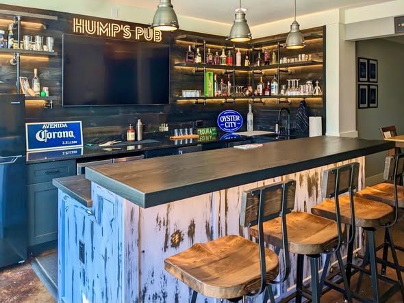 Bar with black countertop, wooden stools, shelves of bottles, and a TV behind the counter