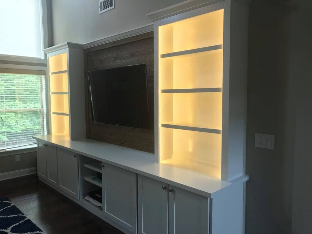 Modern living room wall unit with a TV, illuminated shelves, and white cabinetry by a window