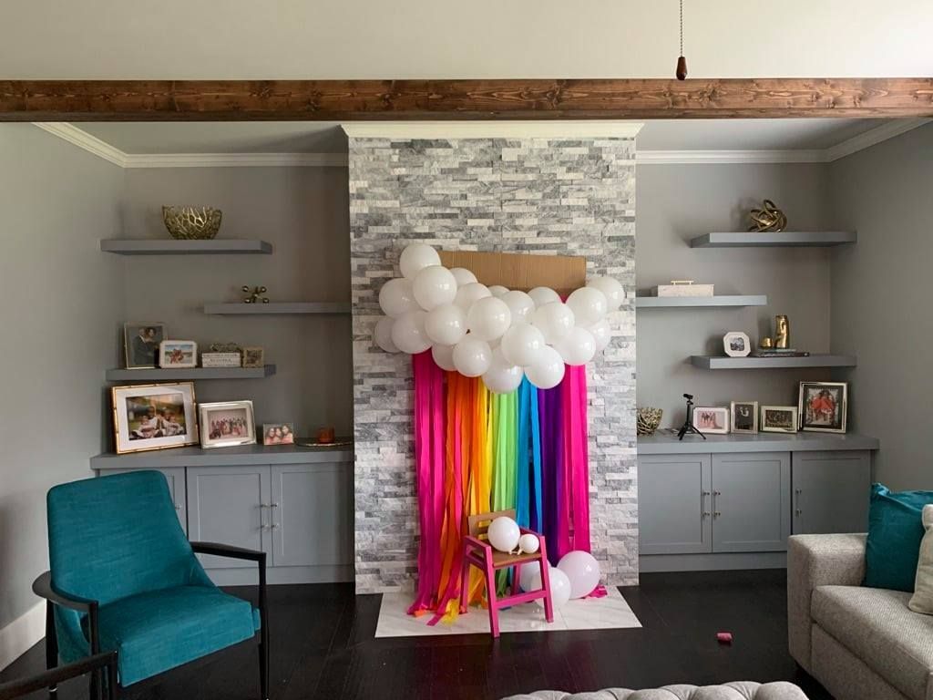 Living room party setup with rainbow backdrop, white balloons, and pink chair in front of a stone fireplace