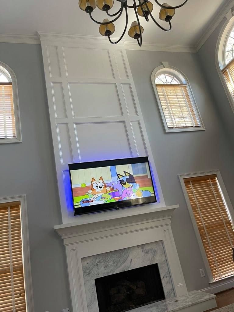 Living room with TV above fireplace showing a colorful cartoon, tall windows, and a chandelier on a high ceiling