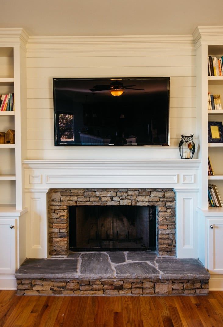 Living room fireplace with mounted TV, white mantel, stone surround, and built-in shelves on both sides