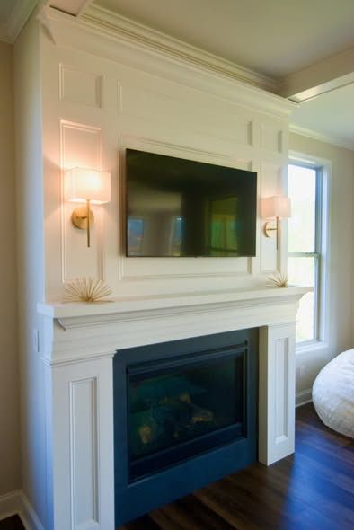White fireplace with mounted TV, wall sconces, and a blue electric fire insert in a bright living room.