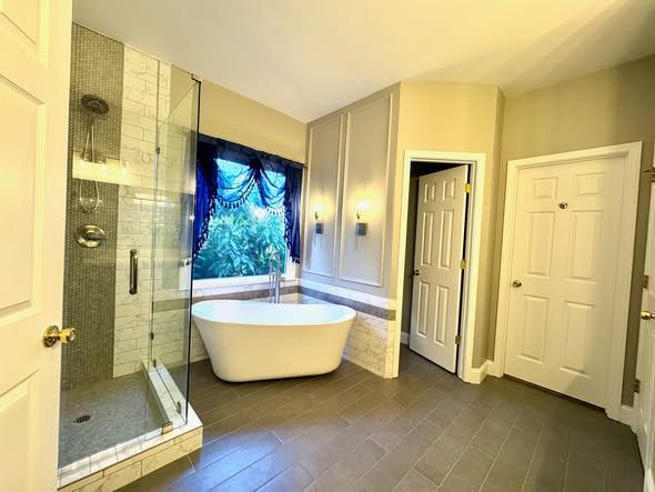 Modern bathroom with freestanding tub, glass shower, and gray tile floor overlooking a window.