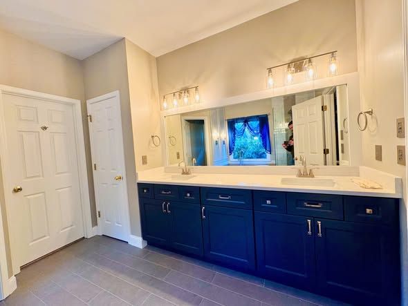 Bathroom vanity with dark blue cabinets, large mirror, and lit wall sconces in a beige room
