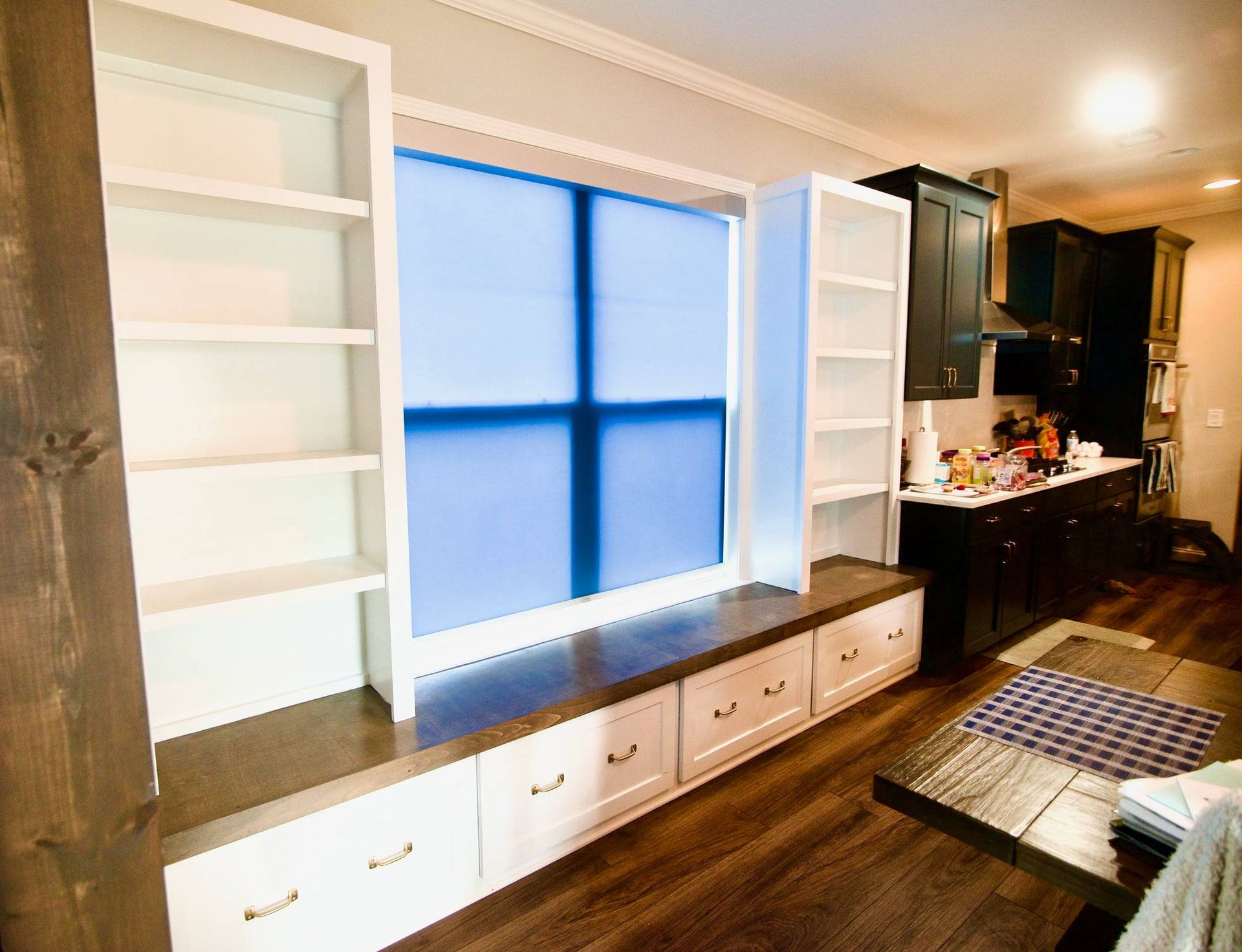 Built-in window nook with white shelves, blue blinds, wood bench, and kitchen area beside it