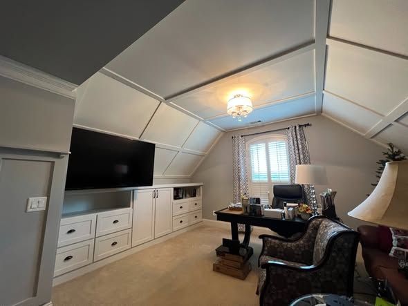 Cozy attic office with sloped ceiling, desk, TV, and armchair near a window with blinds