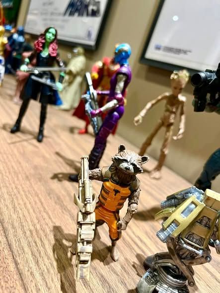 Superhero action figures posed on a tabletop, including Rocket Raccoon in an orange suit in the foreground.