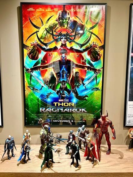 Thor: Ragnarok poster above a lineup of action figures on a wooden shelf.