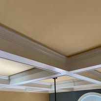 Ceiling with white cross beams and recessed tan panels above a room with a hanging black pendant light.