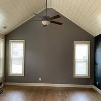 Empty room with vaulted white ceiling, gray accent wall, two narrow windows, and a ceiling fan.