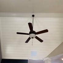 Ceiling fan with dark blades and light fixture on a white shiplap ceiling.