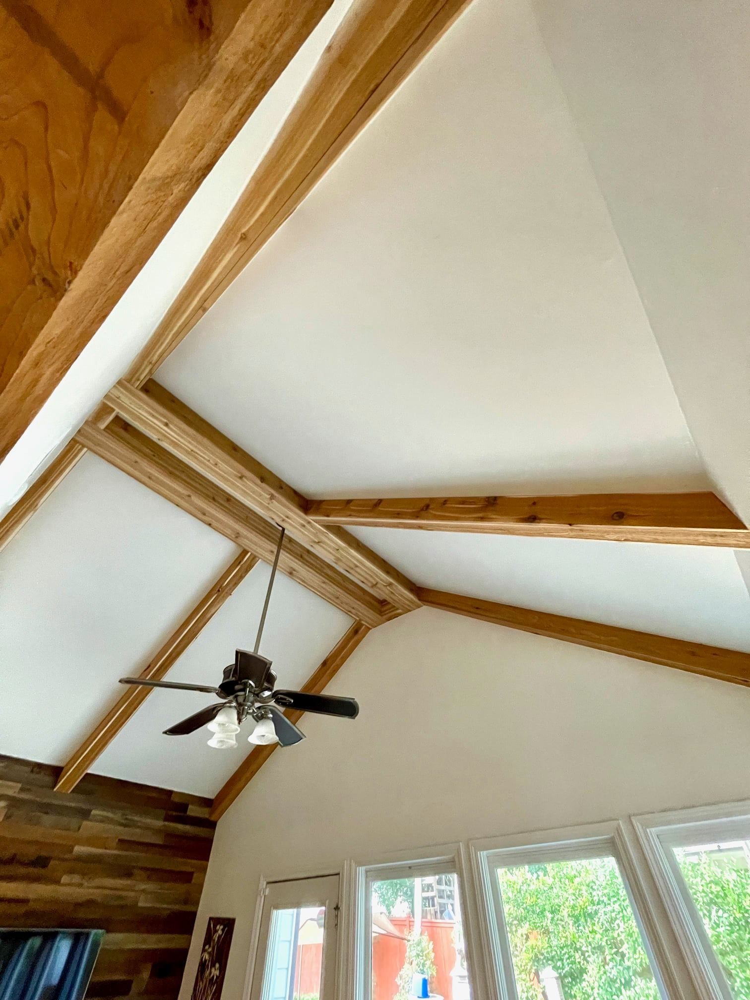 Vaulted ceiling with exposed wooden beams, a ceiling fan, and windows in a bright room