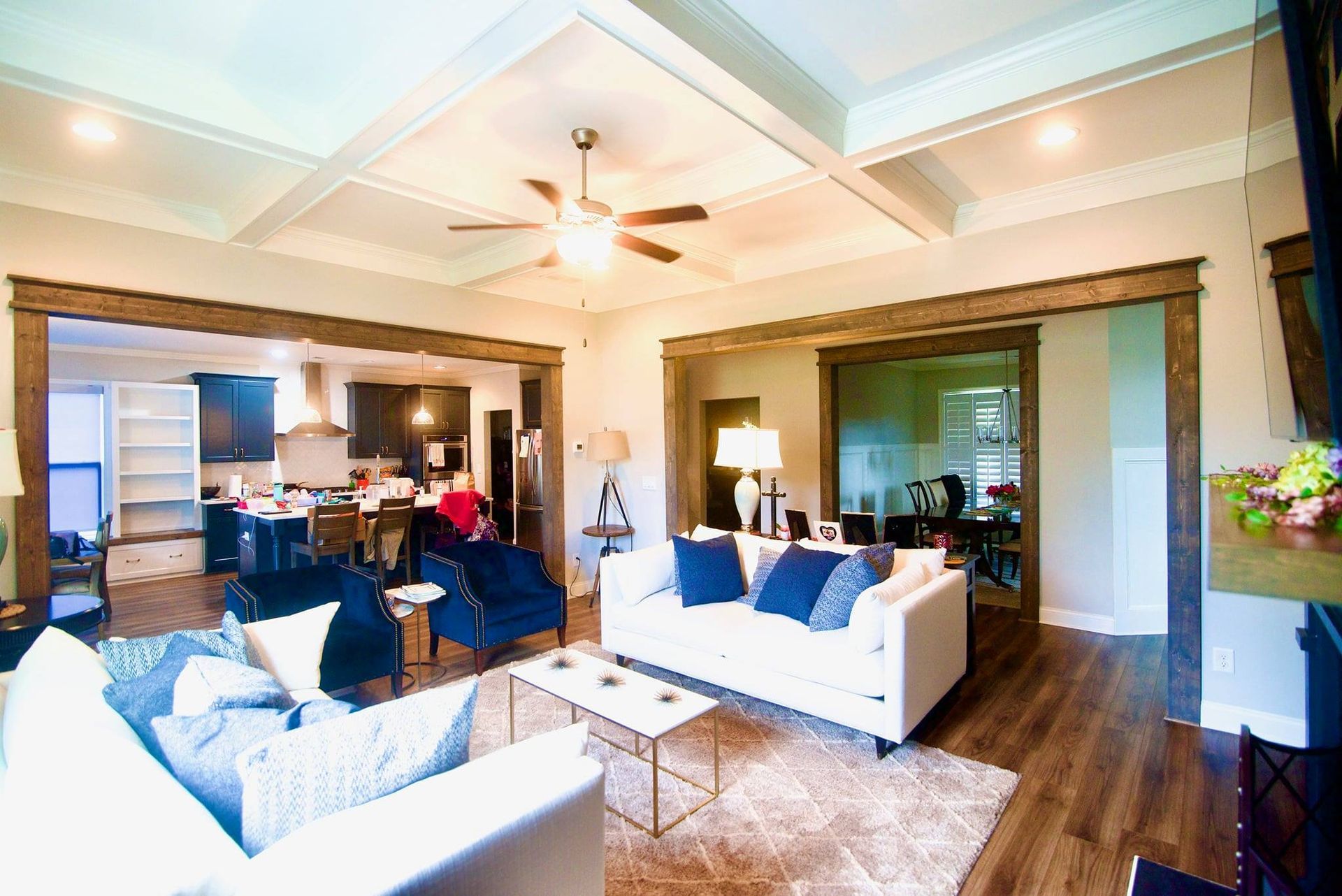 Bright open living room with white sofas, blue pillows, and a ceiling fan opening to a kitchen area.