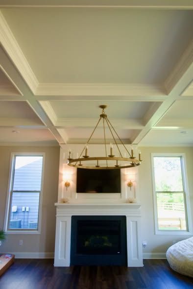 Living room fireplace with white coffered ceiling, large chandelier, and two tall windows on either side