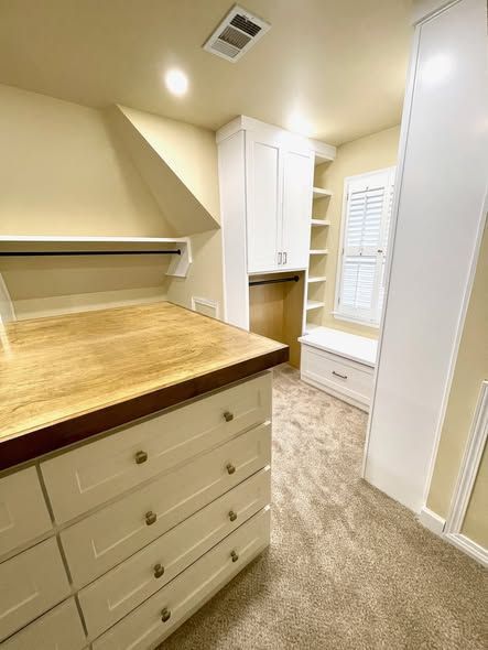 Narrow walk-in closet with white cabinets, wood countertop, shelves, and carpeted floor in warm lighting