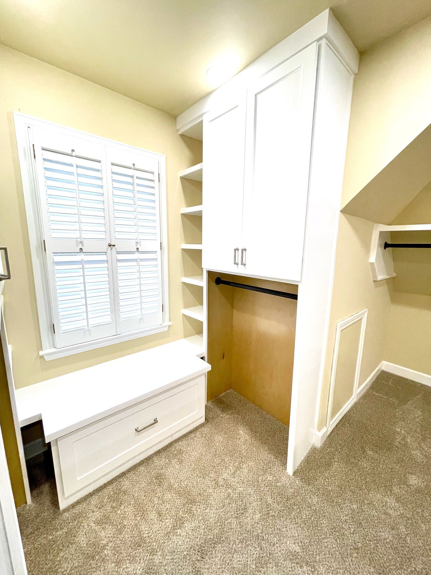 Small beige bedroom nook with window bench, white built-in cabinets, and carpeted floor.