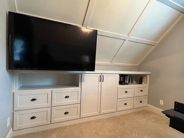 Built-in white TV cabinet with drawers, doors, and a large wall-mounted flat-screen TV in a sloped-ceiling room